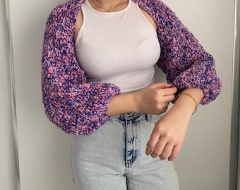 May include: A purple, pink, and blue crochet shrug with long sleeves. The bolero-style sweater has a textured appearance and is worn over a white tank top. The sleeves are gathered at the wrists.