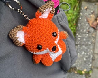 May include: A handmade orange and white crochet fox keychain with black eyes and brown accents. The fox is attached to a silver-colored chain and keyring. The keychain is a cute accessory.