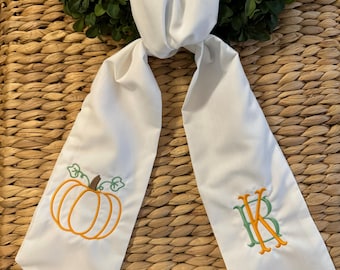 May include: White sash with an embroidered orange pumpkin and green leaves on one end. The other end features embroidered initials in orange and green. The sash is displayed on a woven background with a green wreath.