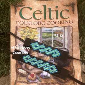 May include: Two beaded bracelets with a black background and blue and green geometric patterns. The bracelets are on top of a book titled "Celtic Folklore Cooking" by Joanne Asala.