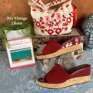 May include: A pair of red cork wedge sandals with a red fabric strap. The sandals are sitting on a white lace tablecloth with a book and a white embroidered bag with red flowers in the background.