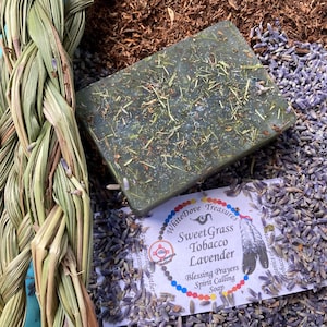 May include: A green soap bar with dried herbs and a label that reads "Sweet Grass Tobacco Lavender". The soap is surrounded by lavender buds, tobacco, and a braided bundle of sweetgrass. The soap is a product of White Dove Treasures.
