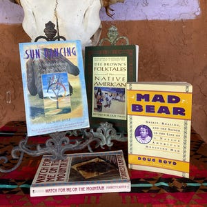 May include: A collection of books with Native American themes, including "Sun Dancing" and "Mad Bear." The books are displayed on a decorative metal stand, with a cow skull in the background. The books have colorful covers.