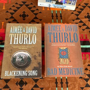 Pode incluir: Dois livros de Aim&eacute;e & David Thurlo s&atilde;o exibidos em uma superf&iacute;cie estampada. O livro &agrave; esquerda &eacute; intitulado "Blackening Song" e apresenta um emblema circular. O livro &agrave; direita &eacute; "Bad Medicine". Um terceiro livro, "The Dark Wind" de Tony Hillerman, &eacute; parcialmente vis&iacute;vel.