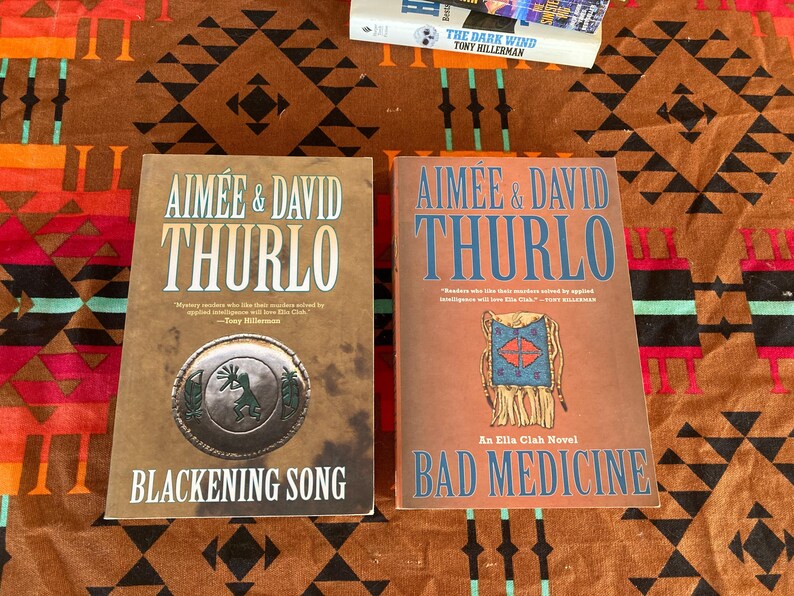 Pode incluir: Dois livros de Aim&eacute;e & David Thurlo s&atilde;o exibidos em uma superf&iacute;cie estampada. O livro &agrave; esquerda &eacute; intitulado "Blackening Song" e apresenta um emblema circular. O livro &agrave; direita &eacute; "Bad Medicine". Um terceiro livro, "The Dark Wind" de Tony Hillerman, &eacute; parcialmente vis&iacute;vel.