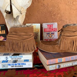 Pode incluir: Botas mocassim de camur&ccedil;a castanha com franjas, exibidas com livros de Tony Hillerman e outros. As botas est&atilde;o sobre uma superf&iacute;cie estampada, com um cr&acirc;nio no fundo.
