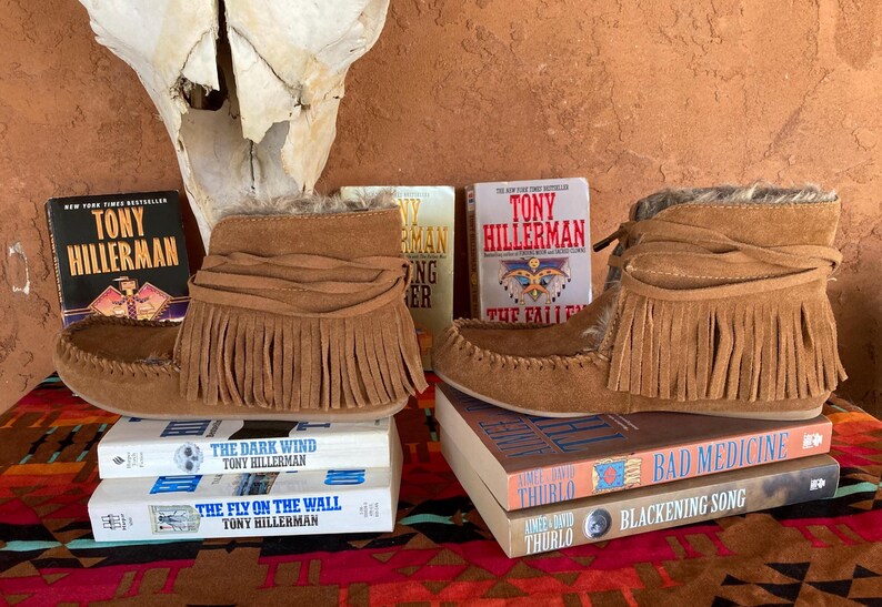 Pode incluir: Botas mocassim de camur&ccedil;a castanha com franjas, exibidas com livros de Tony Hillerman e outros. As botas est&atilde;o sobre uma superf&iacute;cie estampada, com um cr&acirc;nio no fundo.