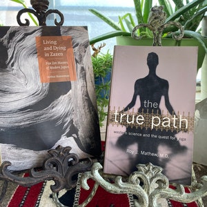 May include: Two books on display, one titled "Living and Dying in Zazen" with a black and white wood grain cover, and the other "the true path" with a silhouette of a person in a yoga pose. Both books are propped up by decorative metal stands.