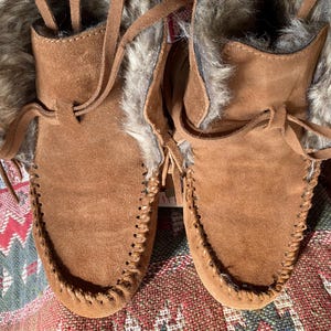 Pode incluir: Um par de botas mocassim de camur&ccedil;a castanha com acabamento em pele sint&eacute;tica. As botas t&ecirc;m um design costurado &agrave; volta da biqueira e um fecho de atacadores em pele. Estas botas s&atilde;o concebidas para o frio.