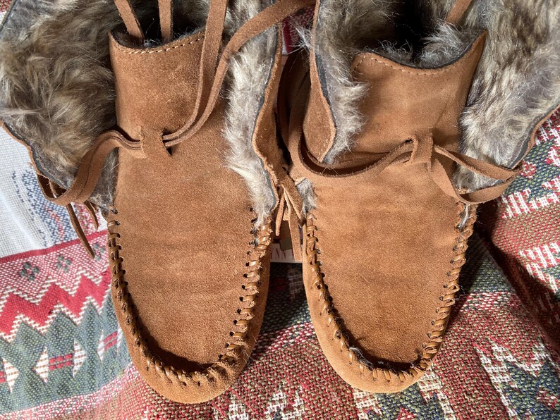 Pode incluir: Um par de botas mocassim de camur&ccedil;a castanha com acabamento em pele sint&eacute;tica. As botas t&ecirc;m um design costurado &agrave; volta da biqueira e um fecho de atacadores em pele. Estas botas s&atilde;o concebidas para o frio.