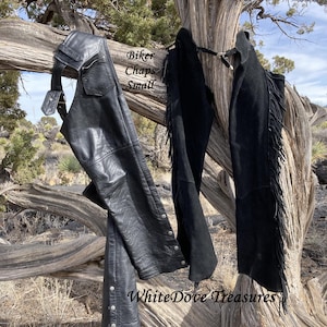 May include: Black leather biker chaps with silver-tone snap closures and a pocket. The chaps are displayed on a tree branch with a pair of black suede chaps with fringe. The text "Biker Chaps Small" is visible.
