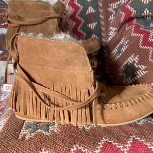 Pode incluir: Botas mocassim de camur&ccedil;a castanha com franjas e la&ccedil;os. As botas t&ecirc;m um aspeto macio e texturizado, colocadas sobre um tecido estampado. A cor castanha &eacute; quente e natural.