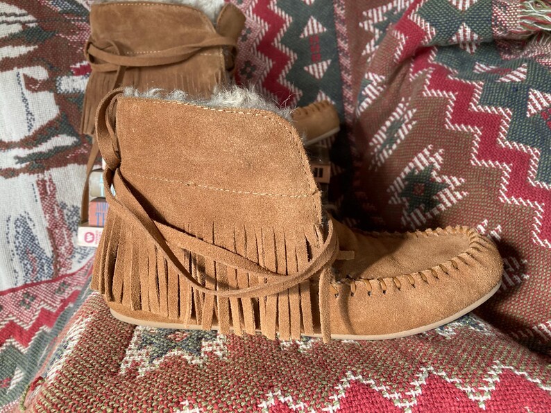 Pode incluir: Botas mocassim de camur&ccedil;a castanha com franjas e la&ccedil;os. As botas t&ecirc;m um aspeto macio e texturizado, colocadas sobre um tecido estampado. A cor castanha &eacute; quente e natural.
