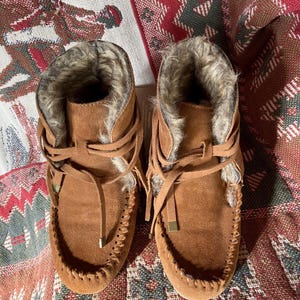 Pode incluir: Um par de botas mocassim de camur&ccedil;a castanha com forro de pele sint&eacute;tica. As botas t&ecirc;m um design com atacadores, costuras decorativas &agrave; volta da biqueira e um pequeno detalhe met&aacute;lico dourado nos atacadores.