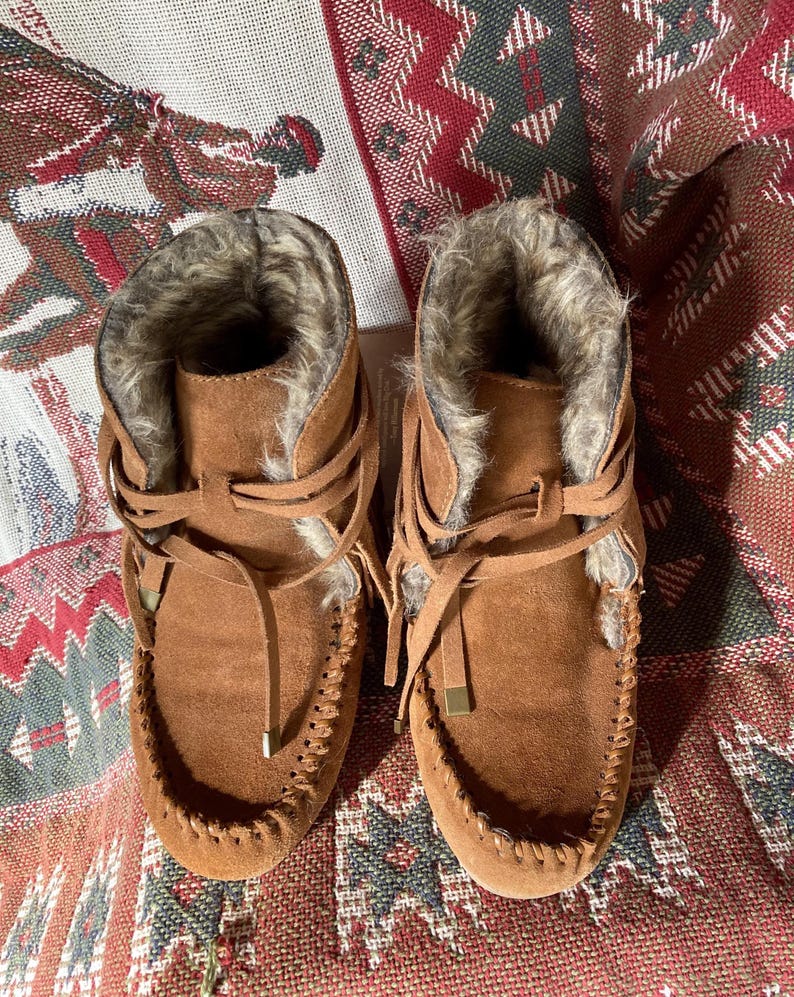 Pode incluir: Um par de botas mocassim de camur&ccedil;a castanha com forro de pele sint&eacute;tica. As botas t&ecirc;m um design com atacadores, costuras decorativas &agrave; volta da biqueira e um pequeno detalhe met&aacute;lico dourado nos atacadores.