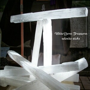 Puede incluir: Un grupo de varillas de selenita blanca dispuestas en forma de cruz. Las varillas son lisas y pulidas, con una apariencia transl&uacute;cida. El texto "White Dove Treasures selenite sticks" es visible en la imagen.
