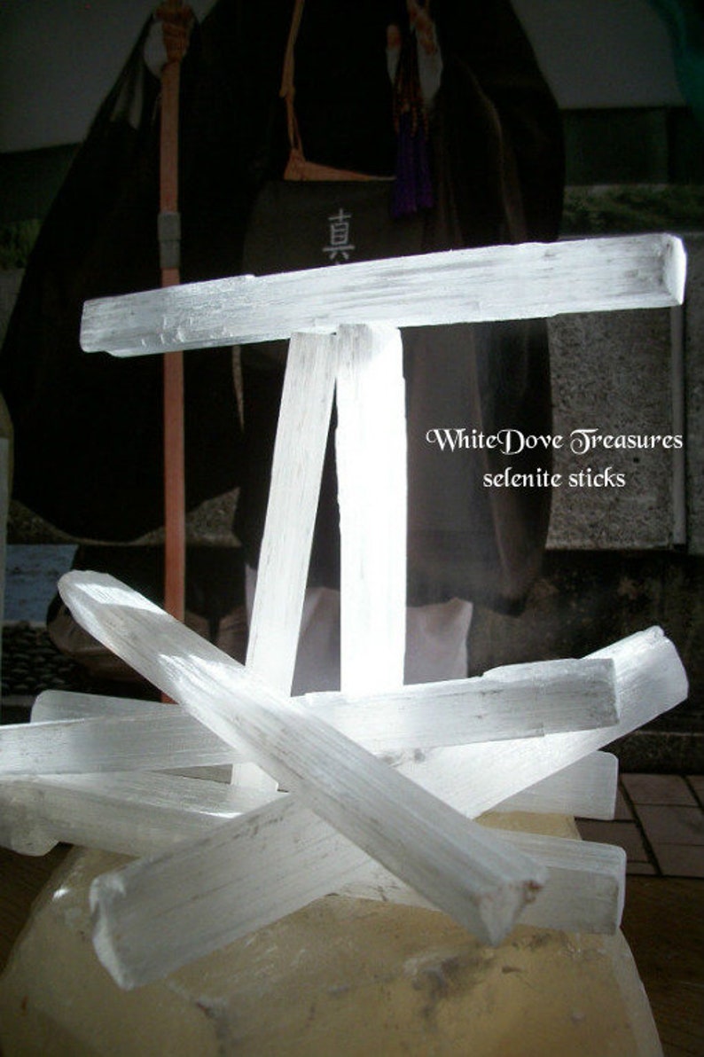 Puede incluir: Un grupo de varillas de selenita blanca dispuestas en forma de cruz. Las varillas son lisas y pulidas, con una apariencia transl&uacute;cida. El texto "White Dove Treasures selenite sticks" es visible en la imagen.