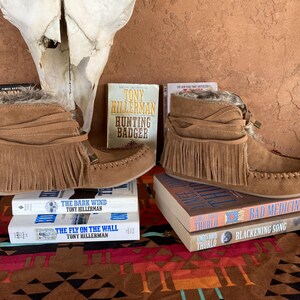 Pode incluir: Botas de camur&ccedil;a castanhas com franjas e acabamento em pele, sobre livros. Os t&iacute;tulos incluem "Hunting Badger" e "The Dark Wind". As botas, estilo mocassim, t&ecirc;m uma faixa decorativa &agrave; volta do tornozelo.