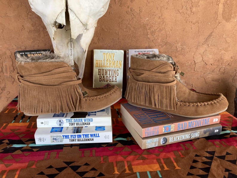 Pode incluir: Botas de camur&ccedil;a castanhas com franjas e acabamento em pele, sobre livros. Os t&iacute;tulos incluem "Hunting Badger" e "The Dark Wind". As botas, estilo mocassim, t&ecirc;m uma faixa decorativa &agrave; volta do tornozelo.