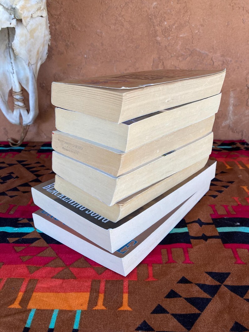 Pode incluir: Uma pilha de sete livros com p&aacute;ginas e capas brancas. Os livros est&atilde;o dispostos numa pilha ligeiramente angulada sobre um tecido estampado. O livro do topo tem uma capa castanha. O livro de baixo tem capas brancas com texto azul.
