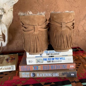 Pode incluir: Um par de botins de camur&ccedil;a castanhos claros com franjas e acabamento em pele. As botas s&atilde;o exibidas numa pilha de livros, incluindo "The Dark Wind" e "Bad Medicine". O fundo apresenta um cr&acirc;nio e um tecido estampado.