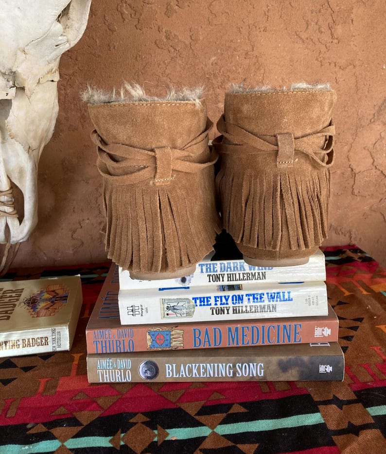 Pode incluir: Um par de botins de camur&ccedil;a castanhos claros com franjas e acabamento em pele. As botas s&atilde;o exibidas numa pilha de livros, incluindo "The Dark Wind" e "Bad Medicine". O fundo apresenta um cr&acirc;nio e um tecido estampado.