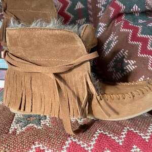 Pode incluir: Botas de tornozelo de camur&ccedil;a castanhas com franjas e bico estilo mocassim. As botas t&ecirc;m um aspeto macio e texturizado e s&atilde;o amarradas com um cord&atilde;o de camur&ccedil;a. As botas est&atilde;o sobre um tecido estampado.