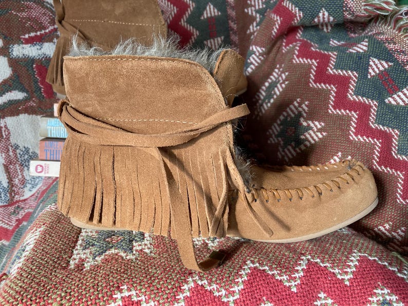 Pode incluir: Botas de tornozelo de camur&ccedil;a castanhas com franjas e bico estilo mocassim. As botas t&ecirc;m um aspeto macio e texturizado e s&atilde;o amarradas com um cord&atilde;o de camur&ccedil;a. As botas est&atilde;o sobre um tecido estampado.