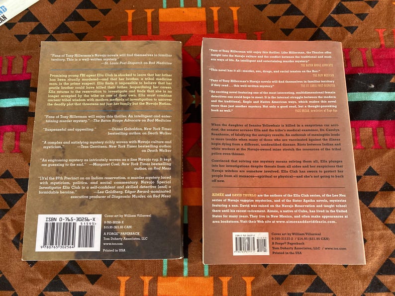 Pode incluir: Dois livros de bolso com capas coloridas s&atilde;o exibidos em um tecido estampado. Os livros apresentam texto e um c&oacute;digo de barras. Um livro &eacute; intitulado "The 7th Precinct on an Indian reservation, a murder mystery laced with mysticism, politics, and social commentary."