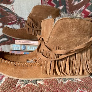 Pode incluir: Um par de botas mocassim de camur&ccedil;a castanhas com franjas. As botas t&ecirc;m um aspeto macio e texturizado e s&atilde;o acentuadas com um la&ccedil;o decorativo. Est&atilde;o sobre uma superf&iacute;cie estampada, com livros ao fundo.