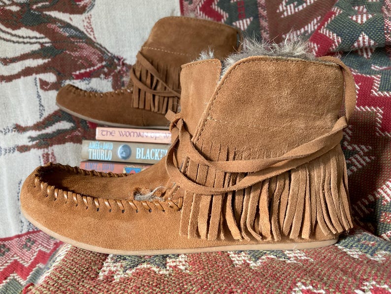 Pode incluir: Um par de botas mocassim de camur&ccedil;a castanhas com franjas. As botas t&ecirc;m um aspeto macio e texturizado e s&atilde;o acentuadas com um la&ccedil;o decorativo. Est&atilde;o sobre uma superf&iacute;cie estampada, com livros ao fundo.