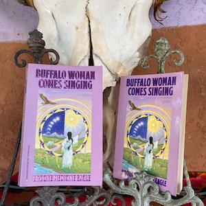 May include: Two copies of the book "Buffalo Woman Comes Singing" by Brooke Medicine Eagle. The book covers are lavender with an illustration of a woman in a white dress, a blue and yellow circle, and an eagle. The books are displayed on ornate metal stands.