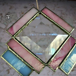 May include: A diamond-shaped stained glass suncatcher with pink and blue glass pieces. The suncatcher is framed in gold metal and has a clear crystal in the center.