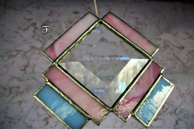 May include: A diamond-shaped stained glass suncatcher with pink and blue glass pieces. The suncatcher is framed in gold metal and has a clear crystal in the center.