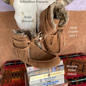 Pode incluir: Mocassins de tornozelo em camur&ccedil;a castanha com franjas e forro de pele sint&eacute;tica, tamanho 7. As botas s&atilde;o exibidas em uma pilha de romances tribais ocidentais, com um cr&acirc;nio de vaca ao fundo. O texto "Ankle Moccasin" &eacute; vis&iacute;vel.