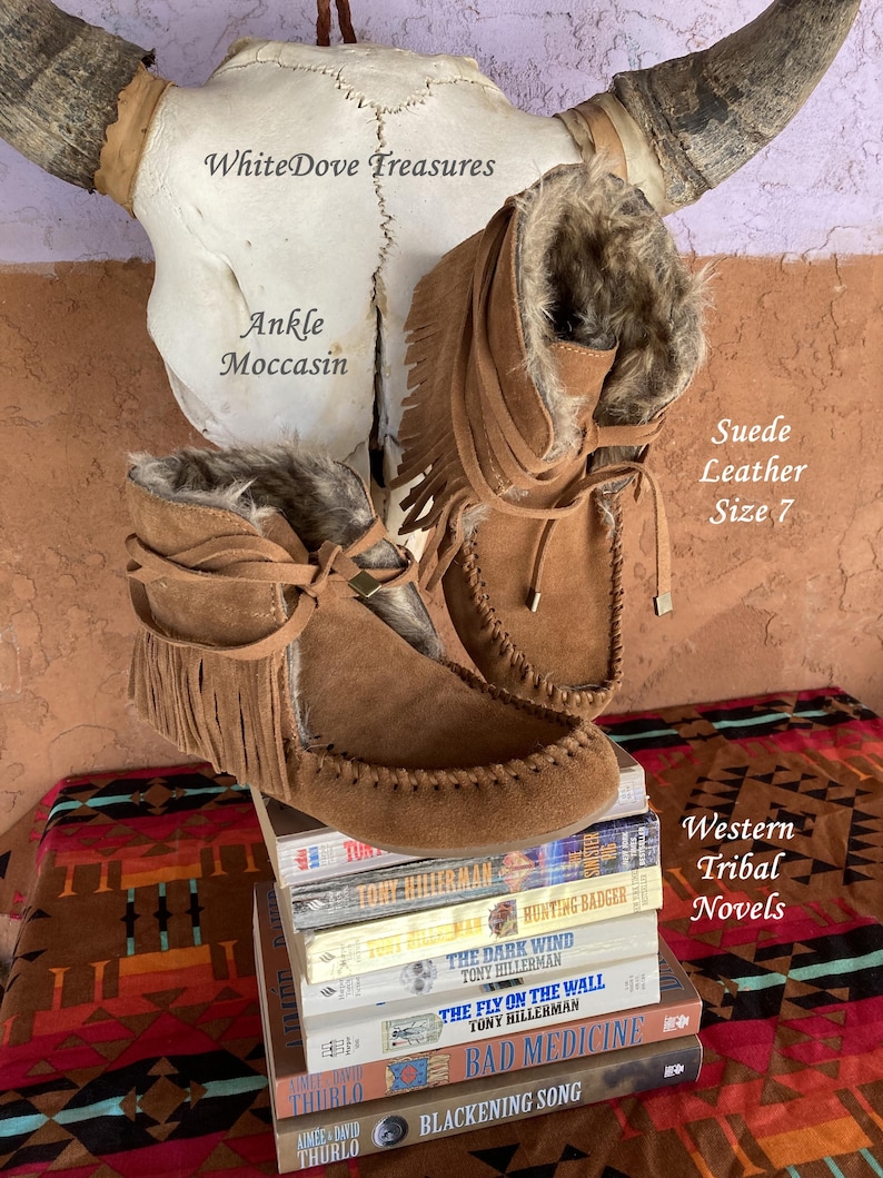 Pode incluir: Mocassins de tornozelo em camur&ccedil;a castanha com franjas e forro de pele sint&eacute;tica, tamanho 7. As botas s&atilde;o exibidas em uma pilha de romances tribais ocidentais, com um cr&acirc;nio de vaca ao fundo. O texto "Ankle Moccasin" &eacute; vis&iacute;vel.
