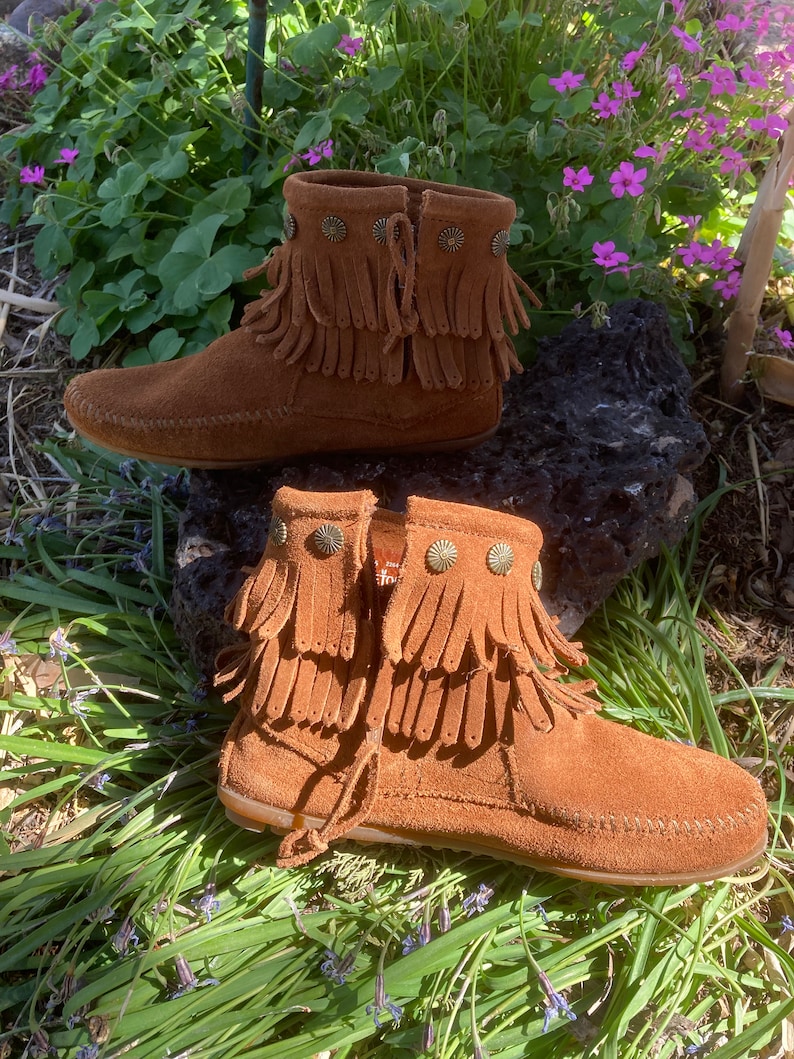 K&ouml;nnte beinhalten: Ein Paar braune Wildlederstiefel mit Fransen und Metallverzierungen. Die Stiefel haben eine spitze Zehenpartie und einen niedrigen Absatz.