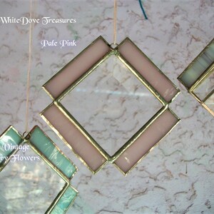 May include: A pale pink diamond-shaped stained glass window hanging from a string. The window has a gold frame and is surrounded by a white background.
