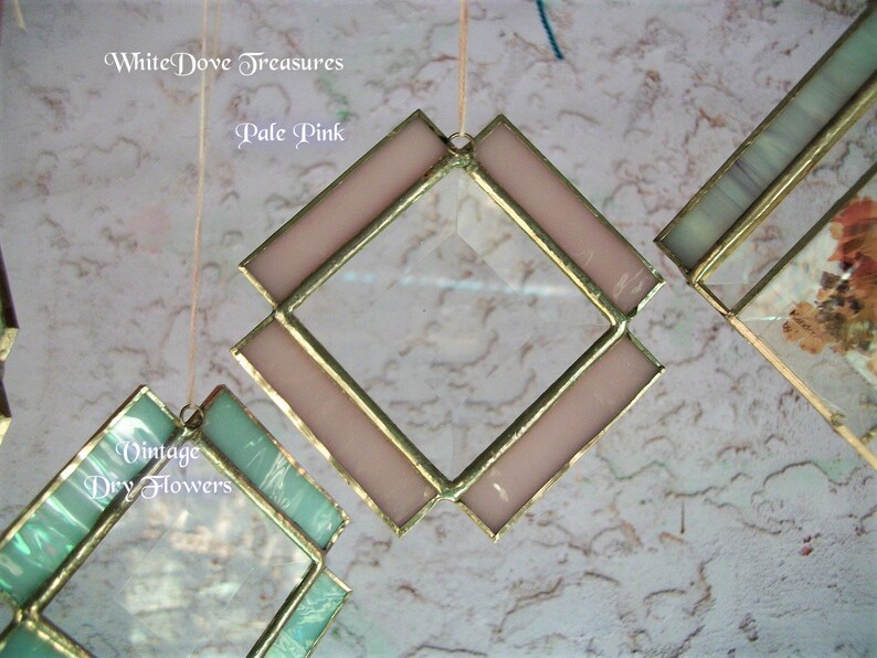 May include: A pale pink diamond-shaped stained glass window hanging from a string. The window has a gold frame and is surrounded by a white background.