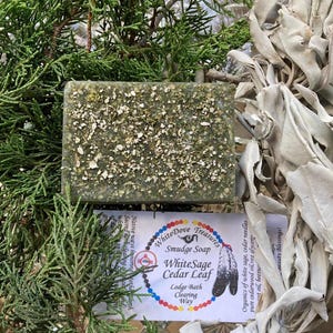 May include: A rectangular green bar of soap with a speckled surface, labeled "WhiteSage Cedar Leaf Smudge Soap." The soap is surrounded by green cedar branches and dried sage leaves. The label includes a logo and text.