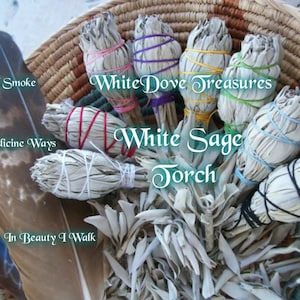 May include: A collection of white sage smudge sticks tied with colorful string in a woven basket. The sticks are labeled "White Dove Treasures White Sage Torch".