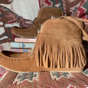 Pode incluir: Botas mocassim de camur&ccedil;a castanha com franjas. As botas de tornozelo t&ecirc;m um design costurado ao redor dos dedos. Uma pilha de livros &eacute; vis&iacute;vel atr&aacute;s das botas. As botas s&atilde;o exibidas em um tecido estampado.