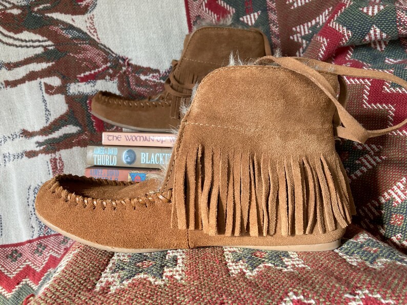 Pode incluir: Botas mocassim de camur&ccedil;a castanha com franjas. As botas de tornozelo t&ecirc;m um design costurado ao redor dos dedos. Uma pilha de livros &eacute; vis&iacute;vel atr&aacute;s das botas. As botas s&atilde;o exibidas em um tecido estampado.