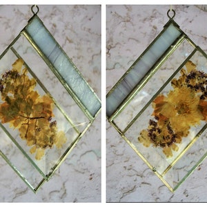 May include: Two square-shaped stained glass ornaments with gold frames. Each ornament features a different pressed flower design. One ornament has a design of dried leaves, while the other has a design of dried flowers.