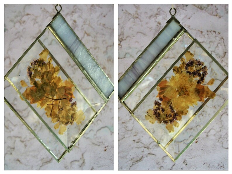 May include: Two square-shaped stained glass ornaments with gold frames. Each ornament features a different pressed flower design. One ornament has a design of dried leaves, while the other has a design of dried flowers.