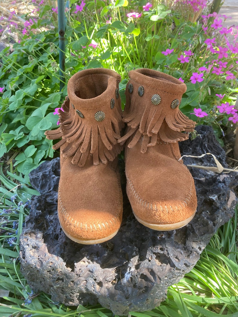 K&ouml;nnte beinhalten: Ein Paar braune Wildlederstiefel mit Fransen und Metallverzierungen. Die Stiefel stehen auf einem Stein mit gr&uuml;nem Gras im Hintergrund.