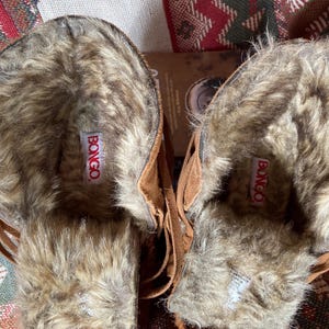 Pode incluir: Um par de botas de camur&ccedil;a castanhas com forro de pele sint&eacute;tica. As botas t&ecirc;m um design com franjas e a marca "BONGO" &eacute; vis&iacute;vel numa etiqueta vermelha dentro de cada bota.