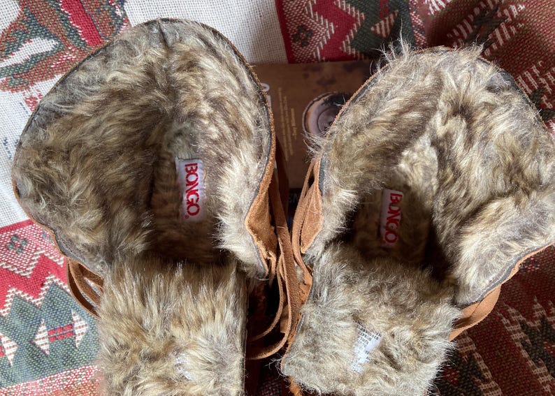 Pode incluir: Um par de botas de camur&ccedil;a castanhas com forro de pele sint&eacute;tica. As botas t&ecirc;m um design com franjas e a marca "BONGO" &eacute; vis&iacute;vel numa etiqueta vermelha dentro de cada bota.
