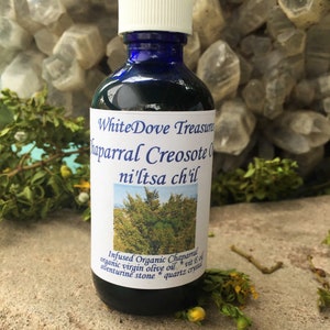 May include: A blue glass bottle with a white lid, labeled "White Dove Treasures Chaparral Creosote Oil ni'ltsa ch'il". The bottle is filled with a dark liquid and is sitting on a light brown surface. The label also says "Infused Organic Chaparral organic virgin olive oil vit E oil aventurine stone quartz crystal".