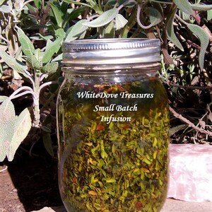 May include: A glass jar filled with green herbs and a clear liquid. The jar has a silver lid and a label that reads "WhiteDove Treasures Small Batch Infusion".